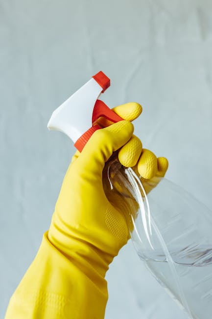 A person wearing yellow rubber cleaning gloves is holding a blue kitchen sponge in one hand and a clear glass spray bottle with a red nozzle and white trigger in the other. The background features a p