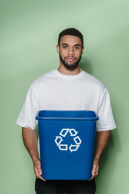 A young man with curly black hair and a beard, wearing a white t-shirt, stands against a light green wall while holding a blue recycling bin with a white recycling symbol on the front. The bin appears