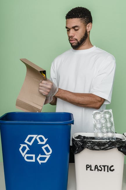 A young man with curly black hair and a beard, wearing a white t-shirt, stands against a light green wall while holding a blue recycling bin with a white recycling symbol on the front. The bin appears