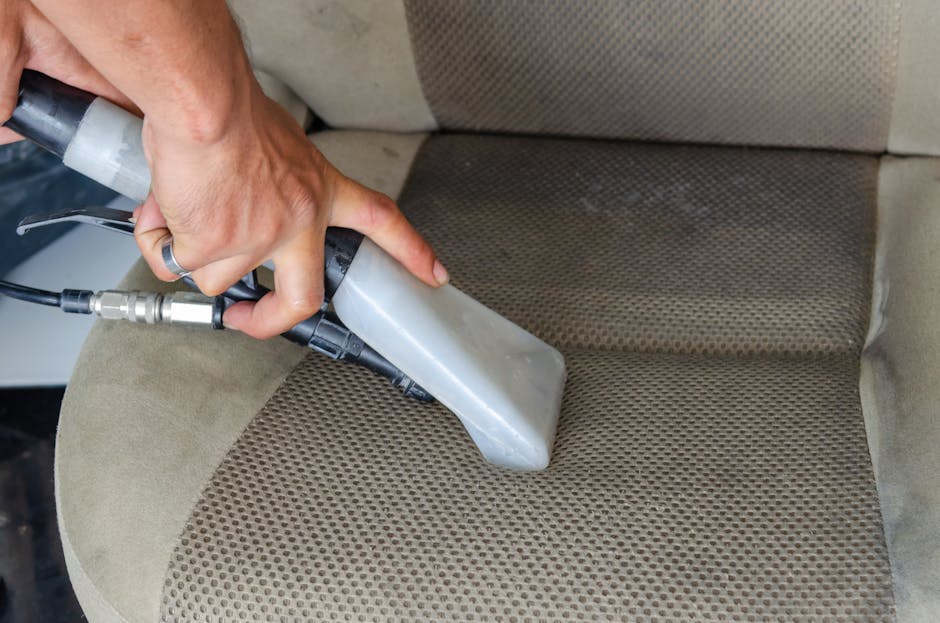 Close-up of a hand vacuuming an olive-green upholstered armchair in a living room, with a dark gray sofa in the background. The upholstery appears clean and well-maintained, with the vacuum cleaner’